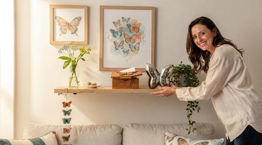 Woman arranging a silver butterfly sculpture beside framed butterfly art, greenery and a wooden keepsake box in a sunlit room.